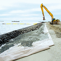An excavator covers installed erosion control mats with the help of riprap revetment on the seashore